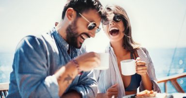 A man and woman sitting on a cruise deck, enjoying coffee and breakfast while laughing together.